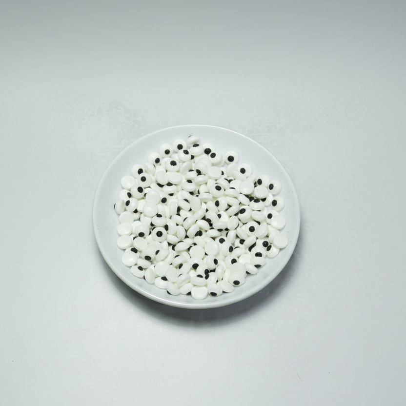 Plato con abundantes ojos comestibles pequeños de azúcar, blancos con pupila negra, para decoración de personajes en repostería. Paquete de 50 gramos de ojitos dulces.